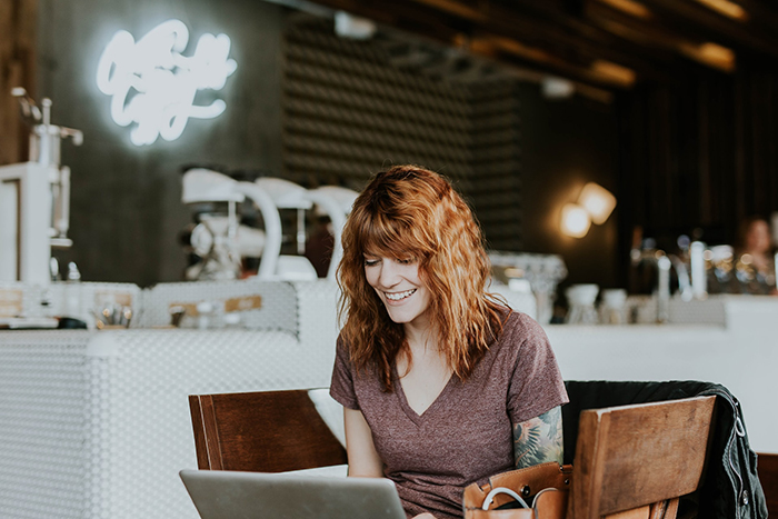 Woman with reddish-brown hair in short sleeve shirt and tattoo on her left arm sitting at a table writing on her laptop. There is a cafe bar in the background.