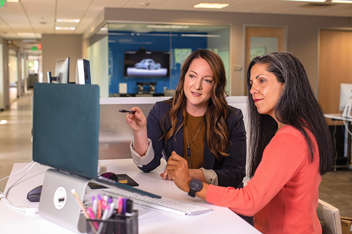 Two women sitting at a desk facing a computer console, only the back of which can be seen. The woman on the left points to the display in front of her, while the woman on the right looks at the same spot.