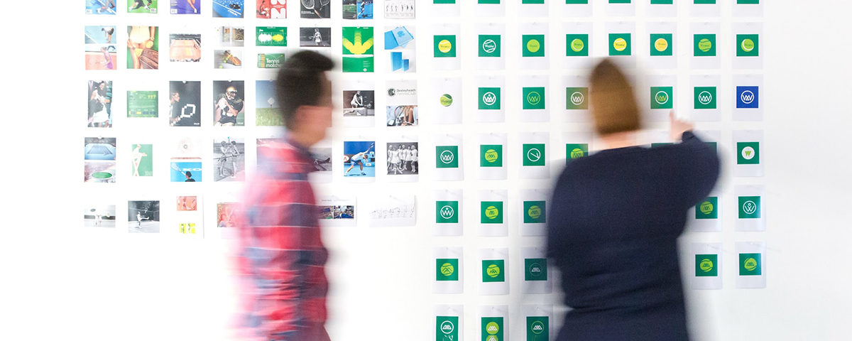 A man in a red and black plaid shirt walking towards a woman in a black top who is looking at a wall that displays different logo and branding options for their real estate company.