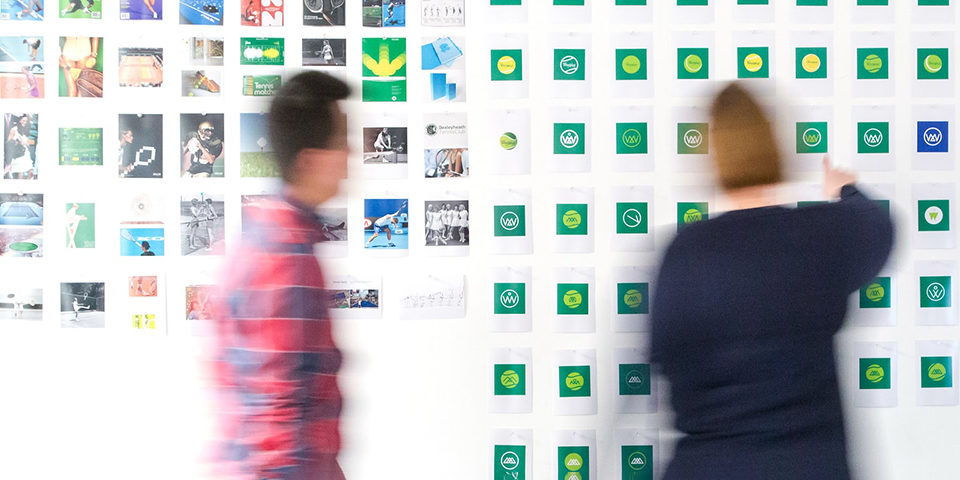 A man in a red and black plaid shirt walking towards a woman in a black top who is looking at a wall that displays different logo and branding options for their real estate company.