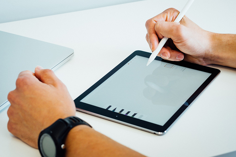 The hands of a man, with a watch on his left wrist, drawing a logo on an iPad that is lying on a flat white desk